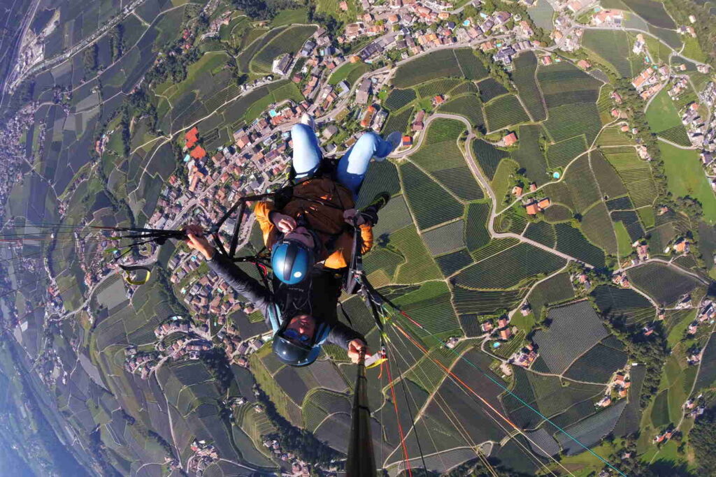 Blick während des Flugs aus der Perspektive vom Gleitschirm aus auf Pilot und Gästin, darunter die Plantagen und Häuser im Tal.