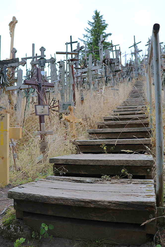Eine Schmale hölzerne Treppe auf die Spitze des Hügels. An den mit wilden Gräsern bewachsenen Seiten des Hügels unzählige Kreuze unterschiedlicher Größe aus verschiedenen Materialien: Eisen, Holz, Stein