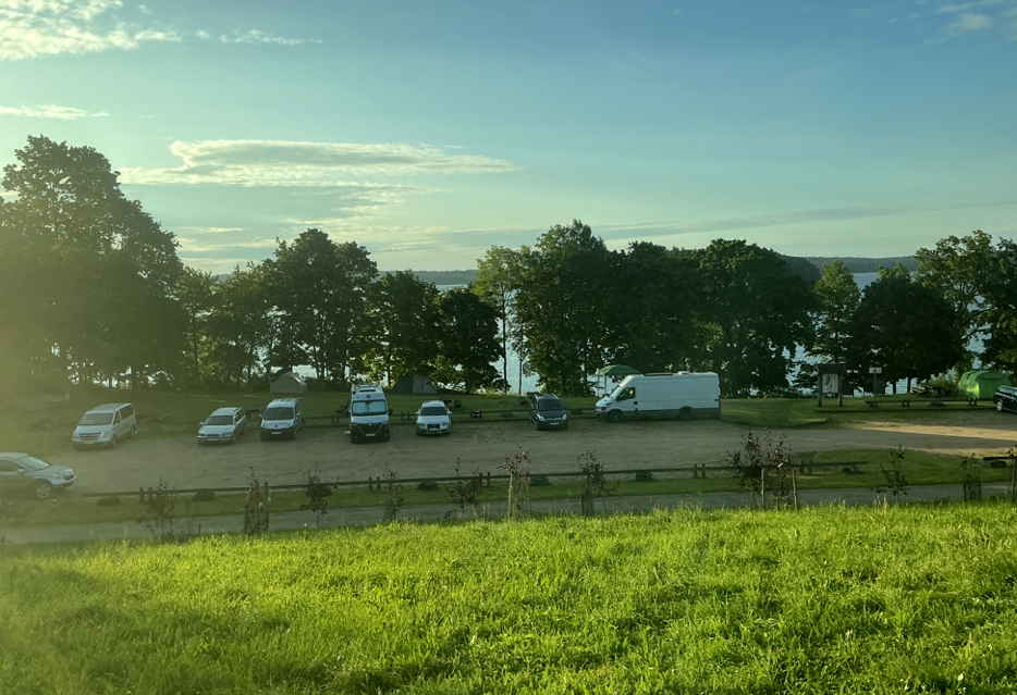 Die erhöhte Wiese für Camper mit Blick auf den Camperparkplatz, eine Baumreihe und den See im Hintergrund.